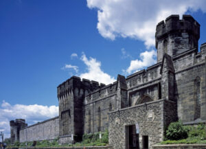 This image shows Eastern State Penitentiary, fortress-like prison with high stone walls, towers, and a Gothic architectural style against a backdrop of a bright blue sky with scattered white clouds.